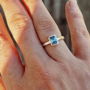Romy Ring with a green radiant cut moissanite in a bezel on a square sterling silver band, worn on the hand